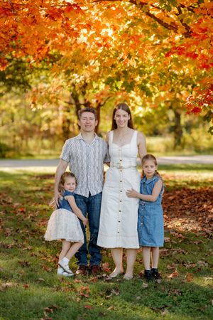 Family of four posing together on a grassy lawn beneath a tree with bright orange autumn leaves.