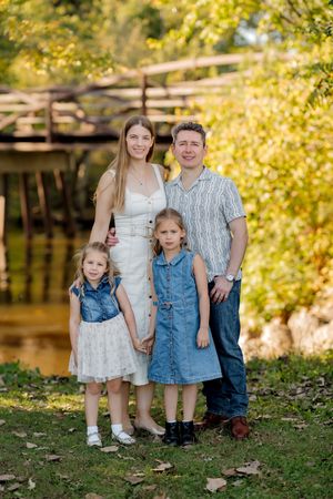 Family of four, a smiling couple with two young girls in denim dresses, pose outdoors near a wooden bridge and autumn foliage.