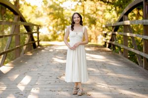Young woman in a flowing white sundress and sandals standing on a sunlit wooden bridge surrounded by green trees.