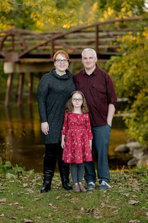 Smiling family of three, a woman, man and young girl in a red floral dress, pose together on a grassy lawn near a wooden bridge and river.