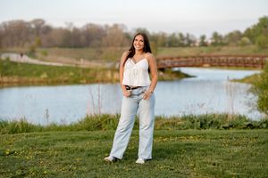 Young woman in white top and light jeans poses on green grass by a scenic pond with a wooden bridge.