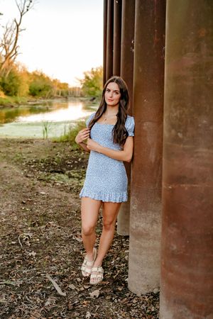 A woman in a blue floral dress poses outdoors near rustic metal columns at sunset.