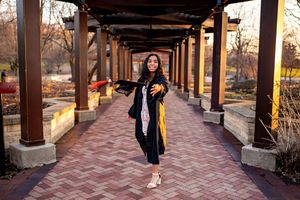A figure in graduation attire with a yellow stole walks happily through a covered outdoor walkway with brick flooring.