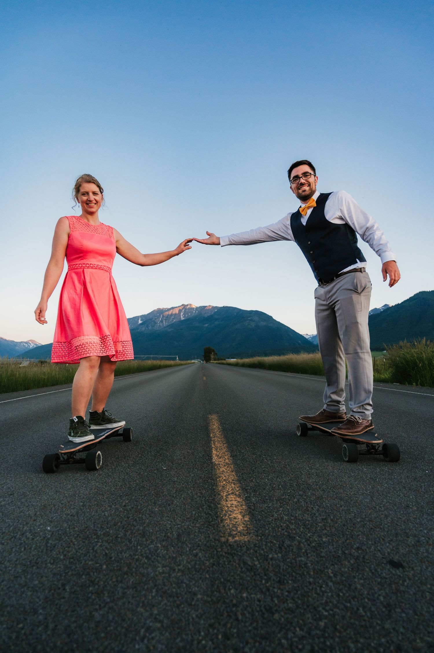 Couple holding hands while skateboarding on empty mountain road at dusk.