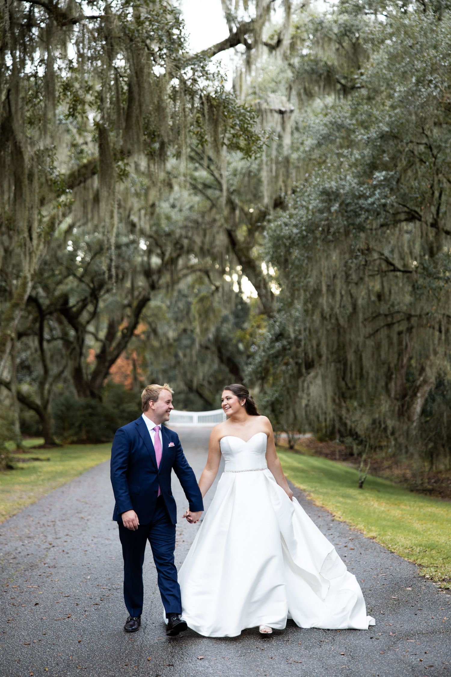 A couple in wedding attire walking together on grass under Spanish moss trees.