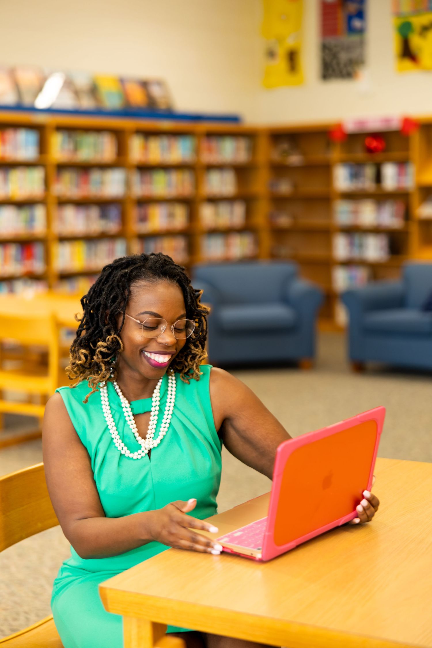 Someone wearing a bright green dress and pearl necklace smiles while using a red laptop in a library setting.