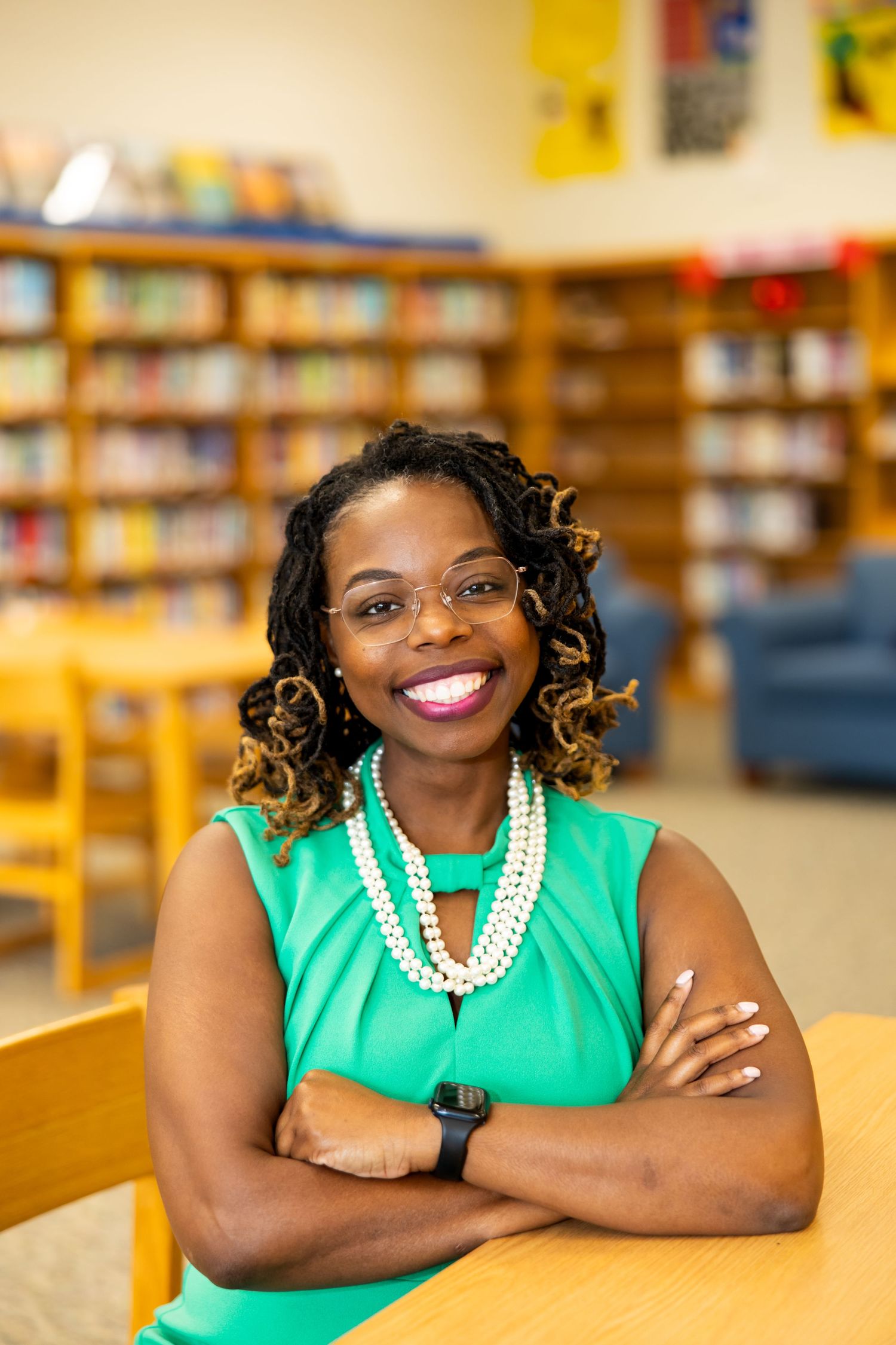 Person in bright green sleeveless top with pearl necklace smiles warmly in library setting.