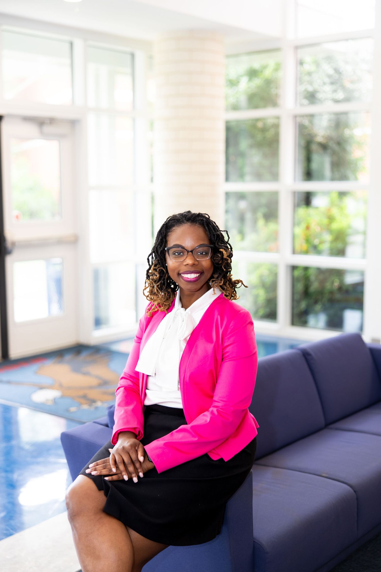 Professional photos showing someone in a pink blazer and white top seated on a navy blue couch in a bright room.