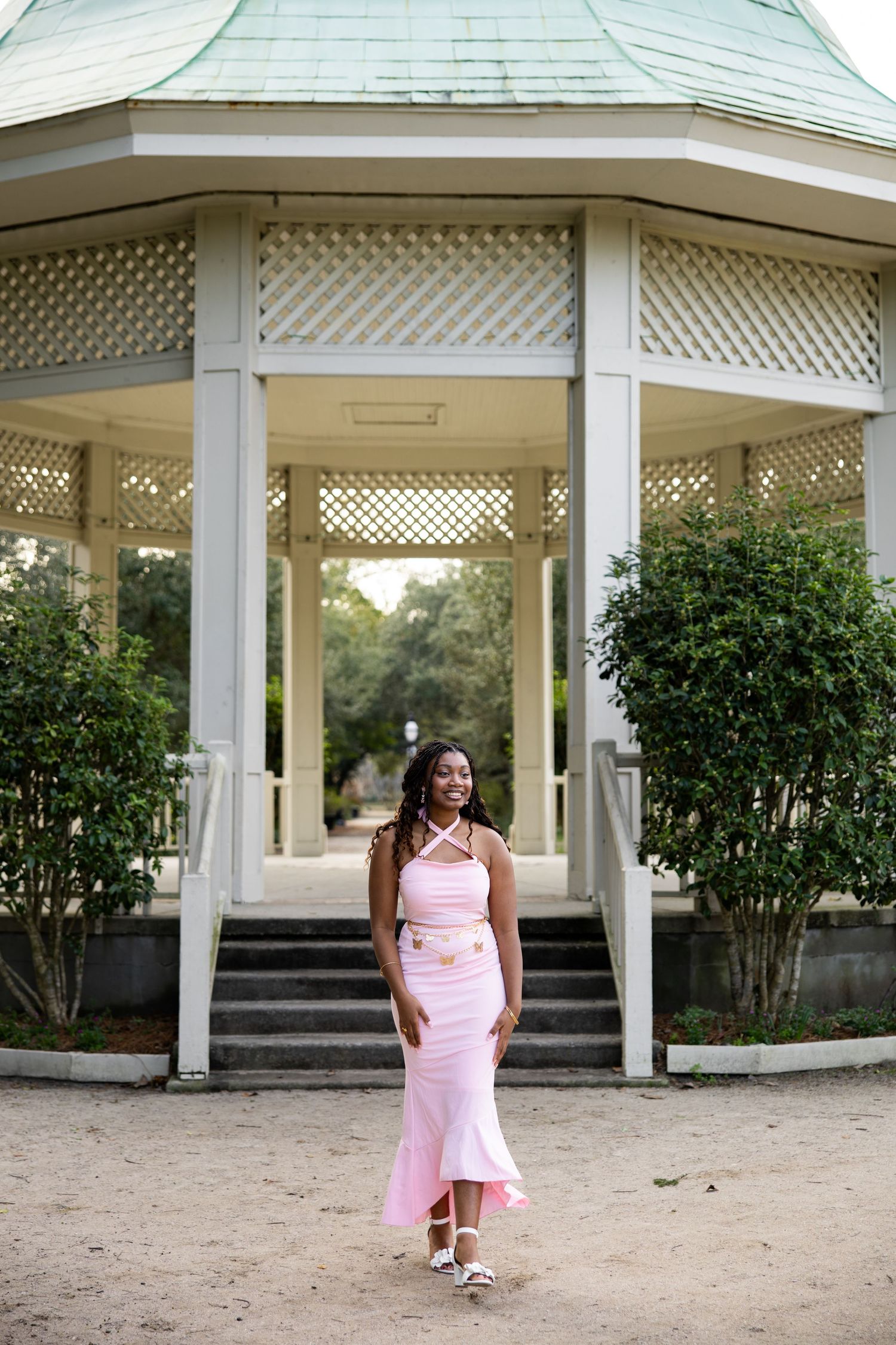 A person in a pink dress stands in front of a white gazebo surrounded by greenery in a garden setting.