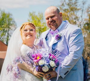 Happy couple poses outdoors on their wedding day with a vibrant purple and white floral bouquet.