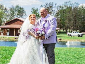 A couple in wedding attire poses on a green lawn with a lake and building in the background.