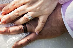 Close up of hands showing gold wedding rings with lace in the background.