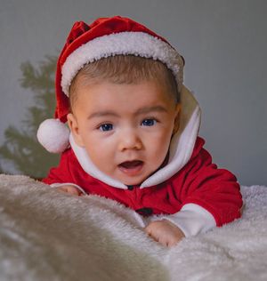 A small baby wearing a red and white Santa outfit and hat lies on soft white fabric with a joyful expression.