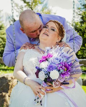 Couple sharing romantic moment on swing with vibrant purple and white floral bouquet.