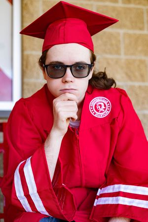 A graduate in a red cap and gown with glasses sits in a thoughtful pose against a brick wall with a school emblem patch.