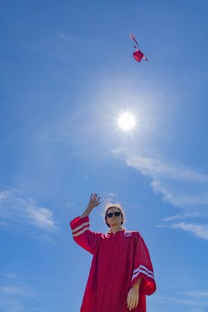 Figure in red graduation gown reaches toward heart-shaped object floating against bright blue sky and sun.