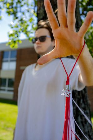Student holding up red graduation tassel against sunny campus backdrop with hand outstretched towards camera.
