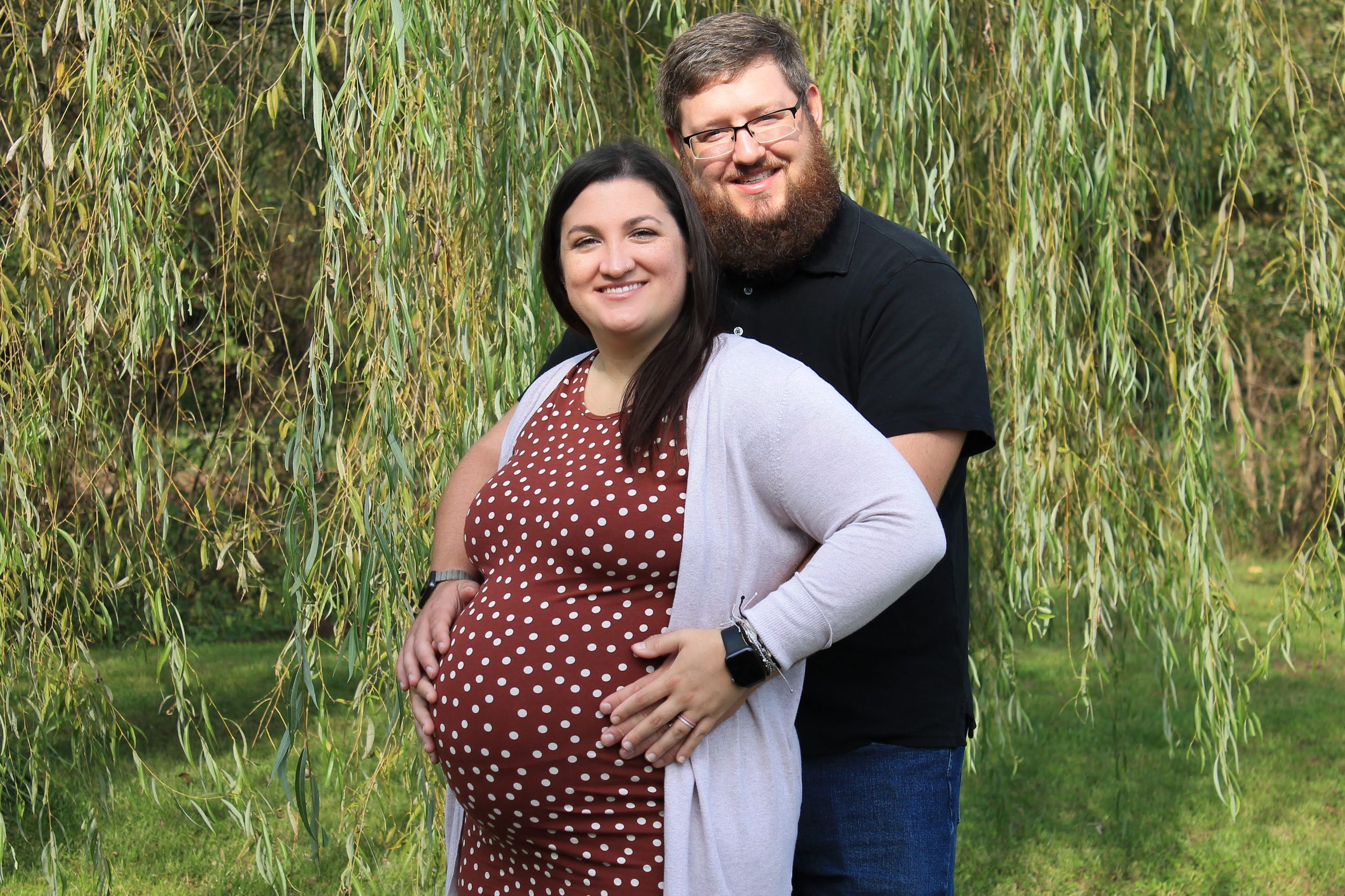A couple poses together under a weeping willow tree, with one wearing a polka dot dress and white cardigan.