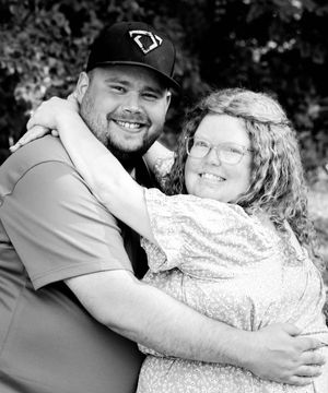 A smiling couple embraces in a black and white portrait photo outdoors with trees in the background.