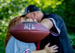 NFL football in foreground with blurred intimate moment in background against green foliage.
