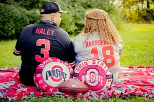 Two Ohio State fans sit on a red blanket wearing custom jerseys and surrounded by OSU football pillows.