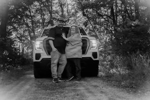 A couple poses in front of a white SUV on a wooded dirt road in black and white photography.