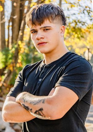 Portrait photo showing someone in a black t-shirt with arm tattoos posing outdoors with autumn trees in the background.