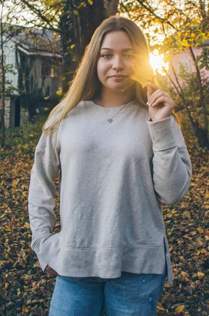 A person in a gray sweatshirt and jeans stands outdoors at sunset with golden sunlight filtering through autumn trees.