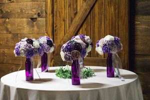 Three purple glass vases holding matching white and purple rose bouquets displayed on a round table with wooden backdrop.