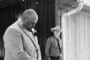 Two people stand outside a building, one in a light suit with boutonniere and another in casual attire in the background.