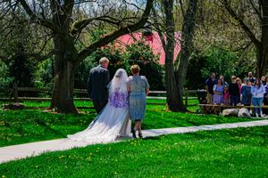 Bride in white dress walks down outdoor garden path surrounded by trees and wedding guests.