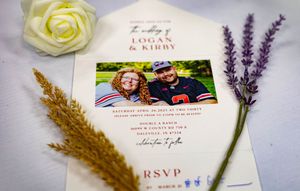 Wedding invitation surrounded by white rose, dried wheat stalk and lavender sprigs on white background.
