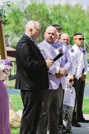 Wedding officiant and groomsmen in formal attire stand at outdoor ceremony altar with purple decor.