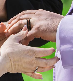 Close-up of hands exchanging wedding rings during a ceremony with one person wearing a light purple sleeve.