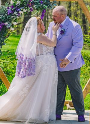 A wedding ceremony takes place under a floral arch decorated with purple flowers and greenery.