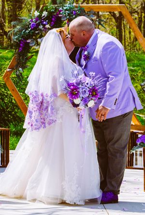 A couple shares a romantic kiss under a purple floral hexagon arch during their outdoor wedding celebration.