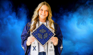 Graduate in navy blue cap and gown opens jacket to reveal decorated graduation cap design against blue smoke background.