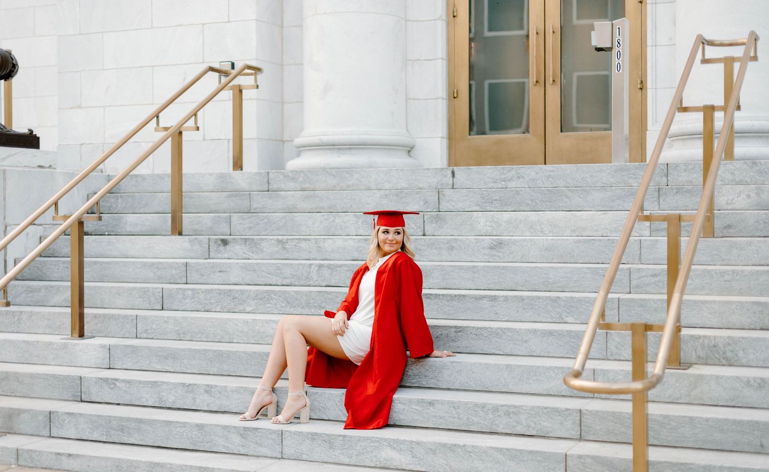 Graduate in bright red cap and gown sitting on white stone steps.