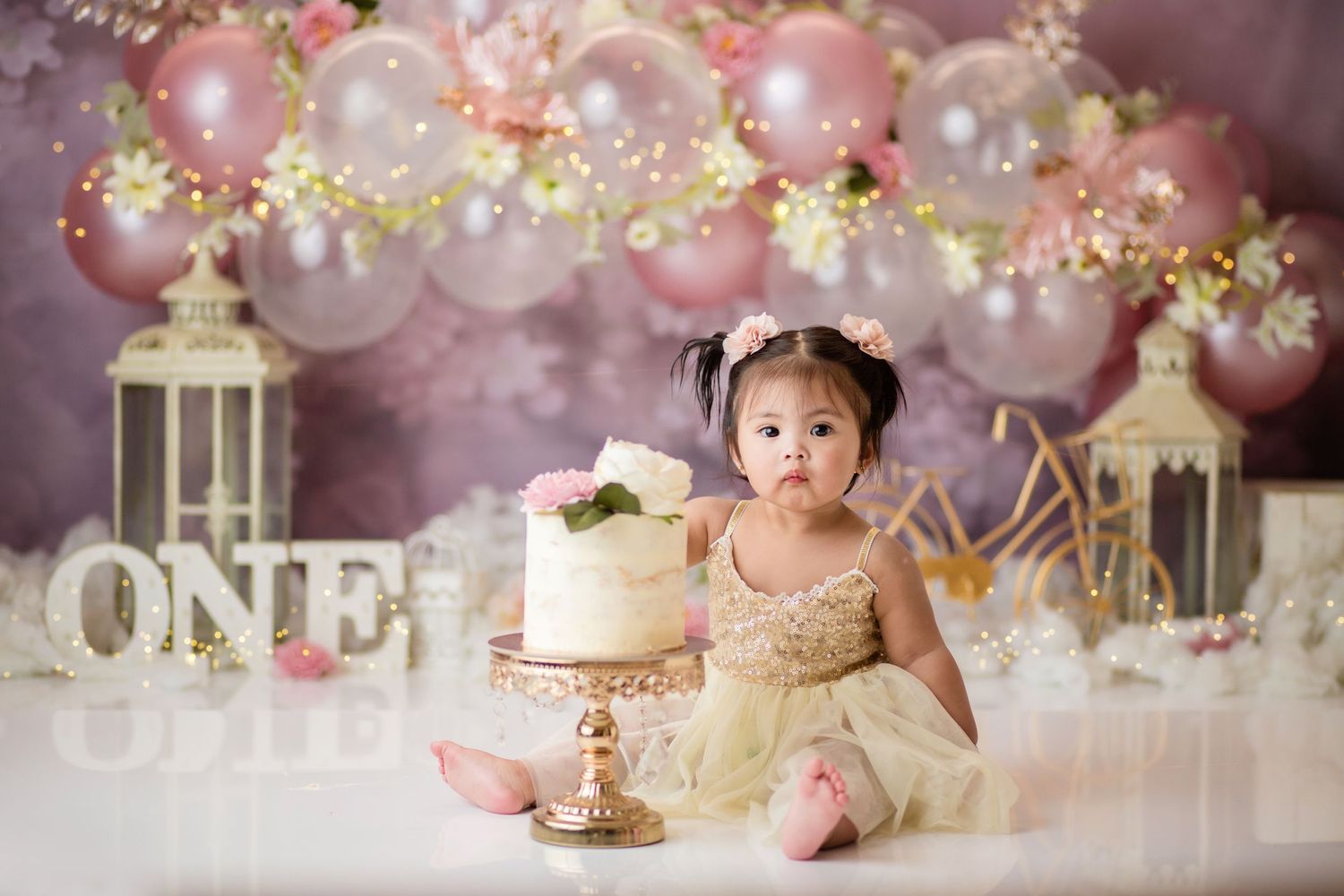 Dreamy pink first birthday setup with lanterns, balloons, and white cake on elegant stand.