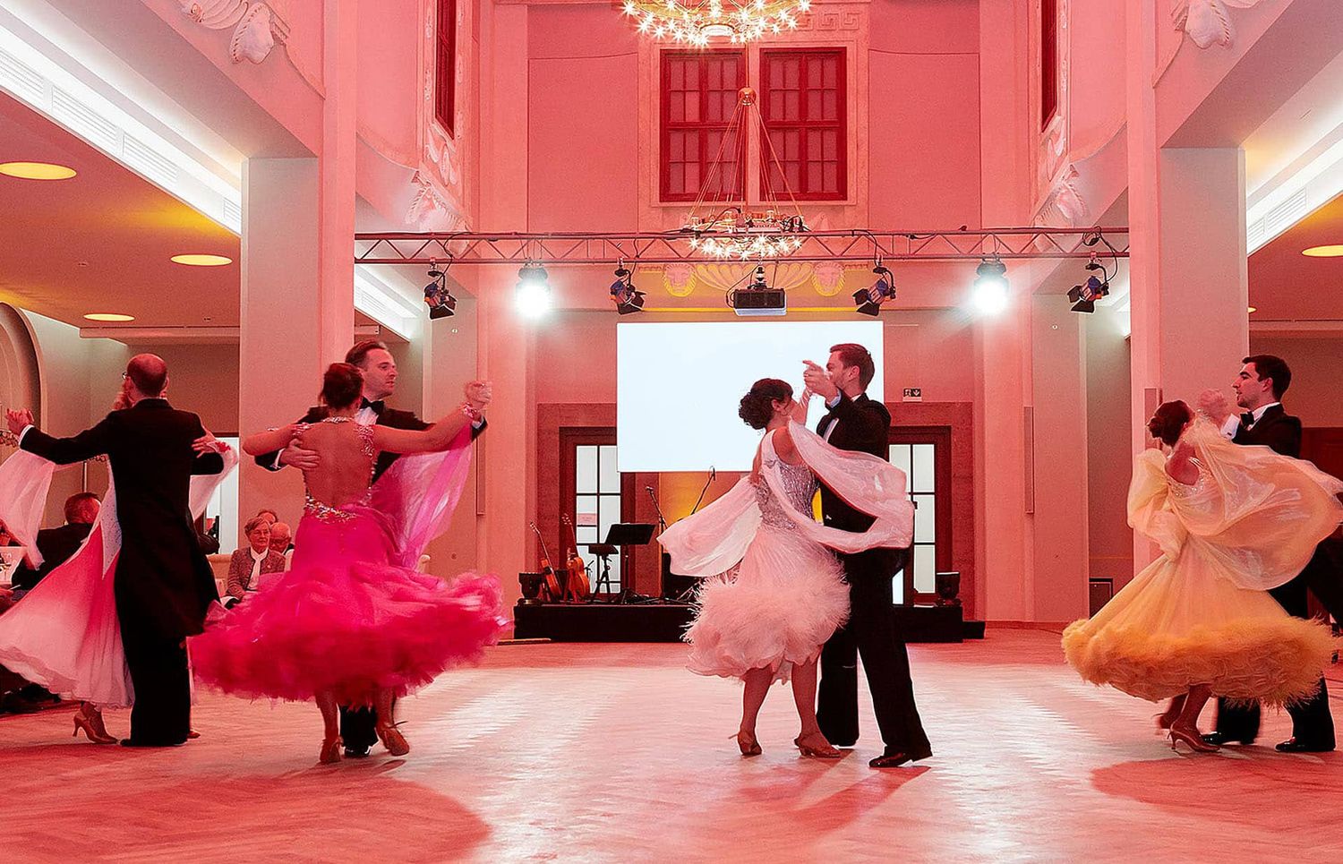 Couples in elegant ballroom attire dance together in a grand pink-hued hall during a formal dance event.