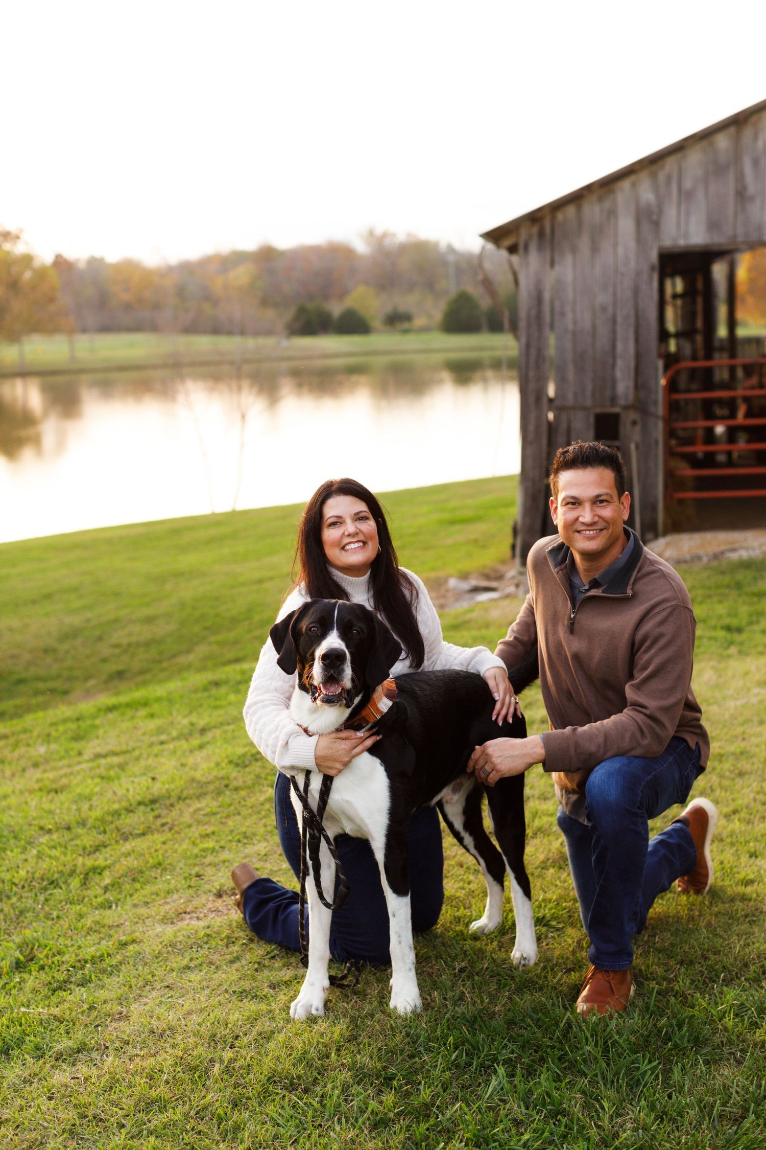 A black and white dog poses with owners by a rustic barn and lake during golden hour.