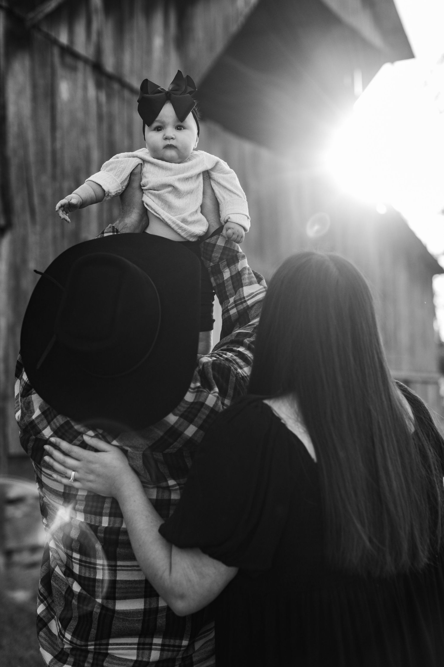 A black and white photo shows a family moment with a toddler being lifted high while wearing a bow against bright sunlight.
