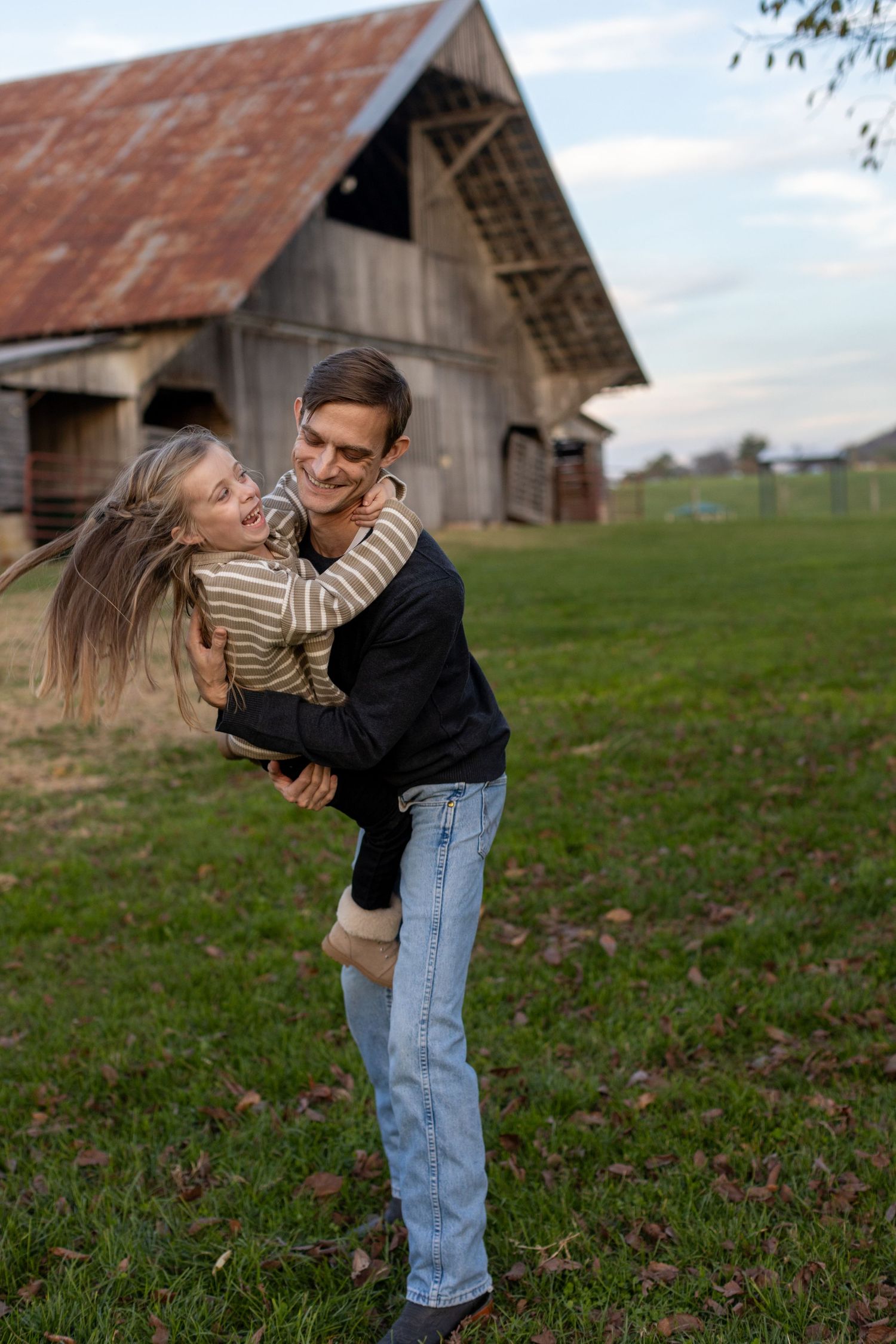 A couple embraces playfully in front of a rustic barn with a tin roof on a grassy countryside landscape at dusk.