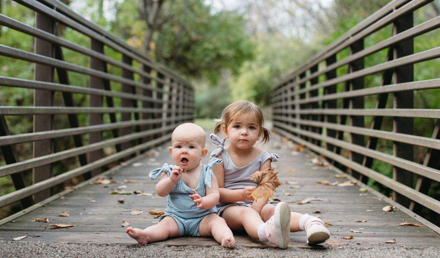 Two small children in casual clothing sit together on a wooden footbridge in a nature setting.