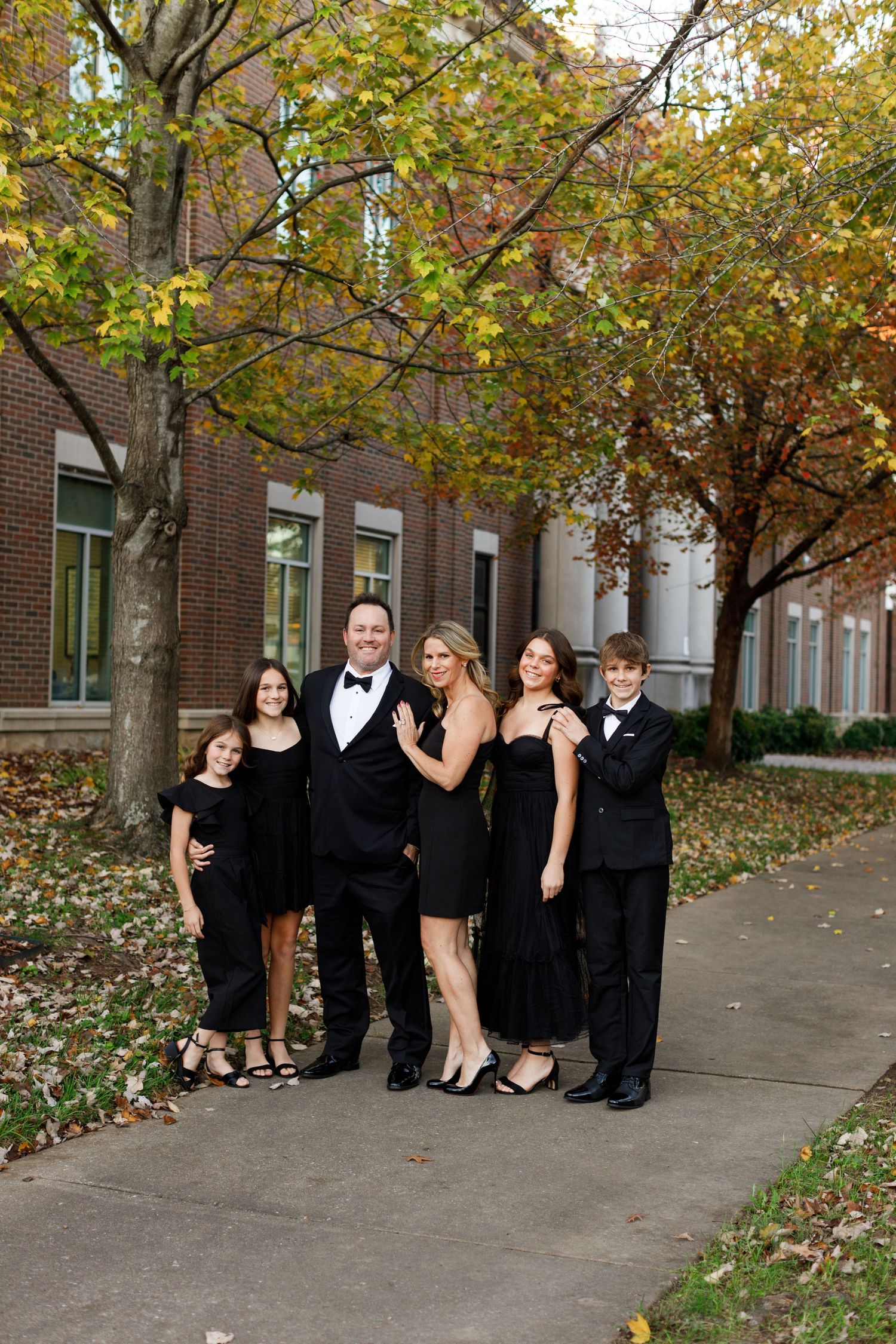 A formal group in black attire poses together on a sidewalk during autumn with trees showing fall colors in the background.