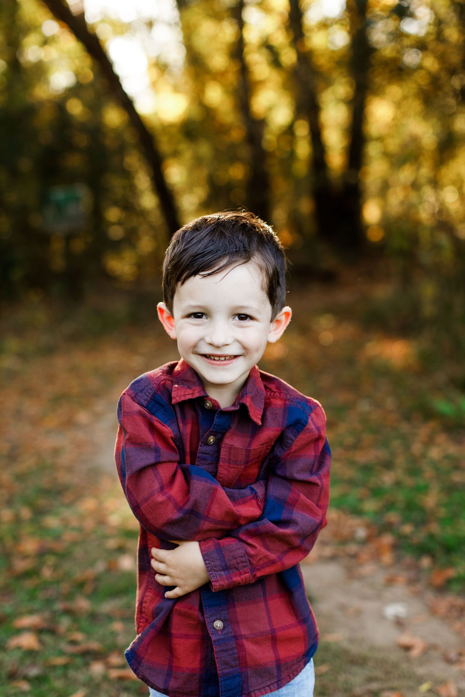 A child in a red and black plaid flannel shirt smiles outdoors during golden hour in an autumn forest setting.