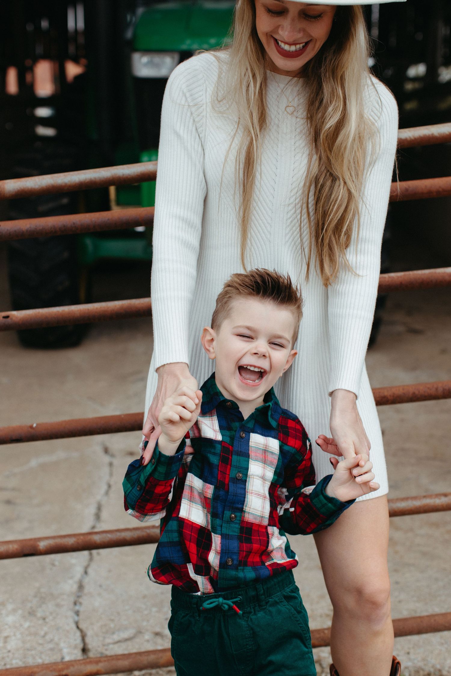 A young child in a plaid shirt and green pants laughs joyfully while standing next to someone in a white sweater.