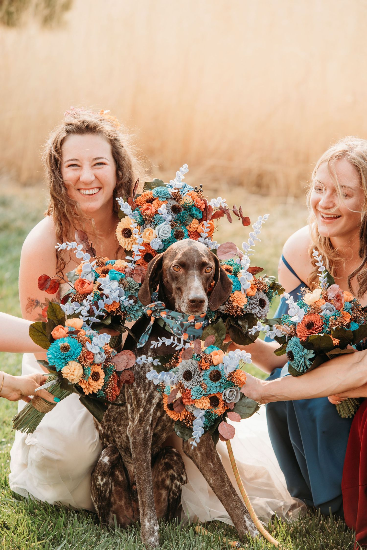 Two attendants pose with a flower-adorned brown dog holding vibrant orange and blue bouquets.
