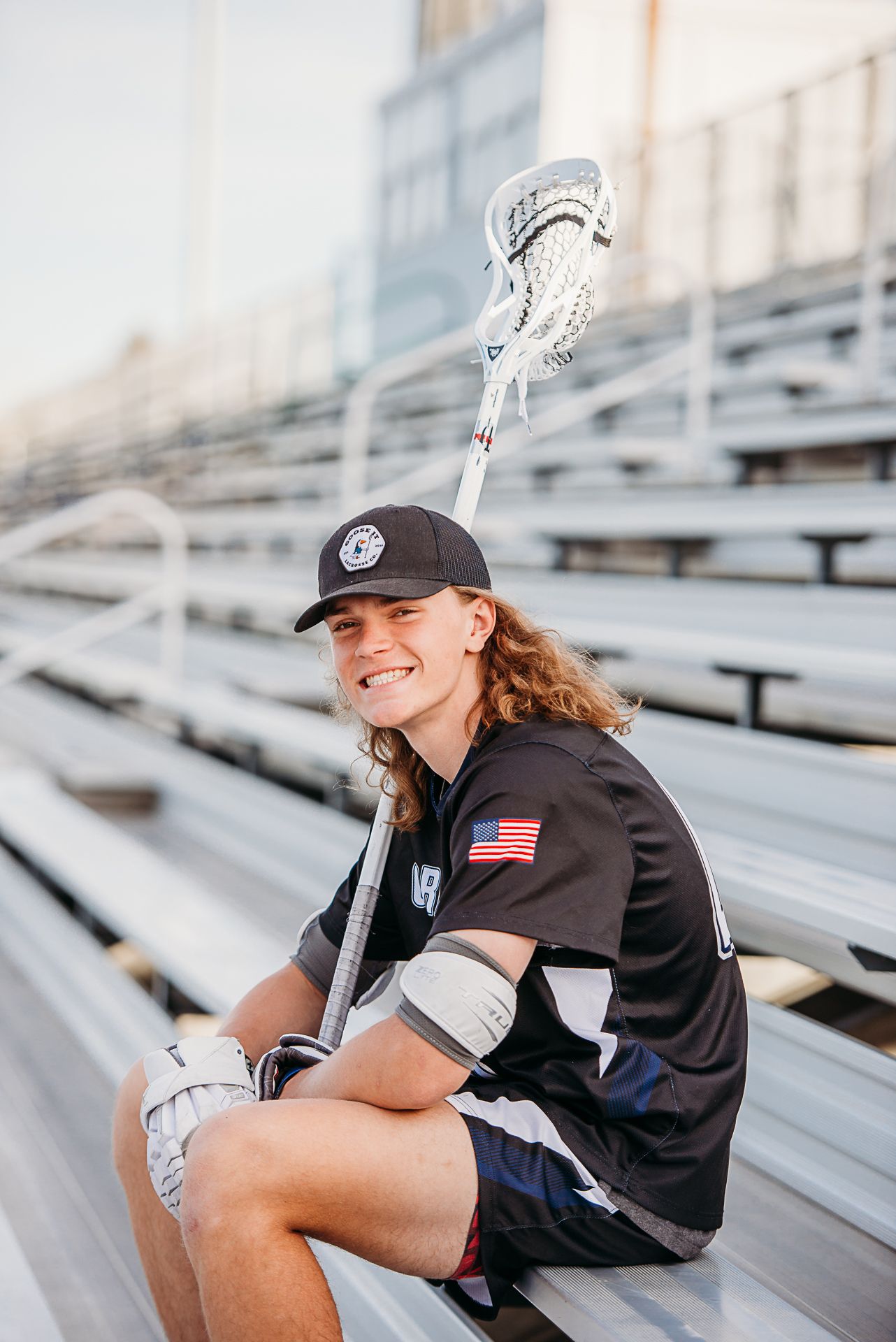 Lacrosse player sitting on stadium bleachers holding a stick in casual pose.