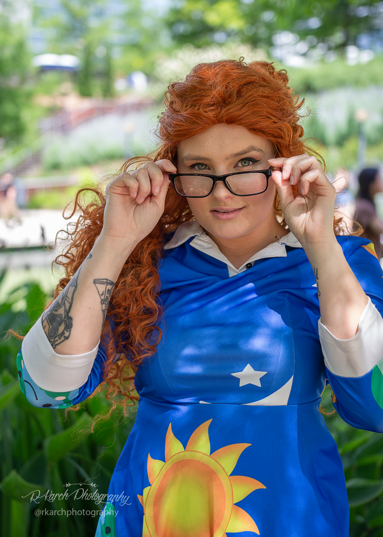 Cosplayer in blue costume with curly red hair adjusting glasses outdoors.